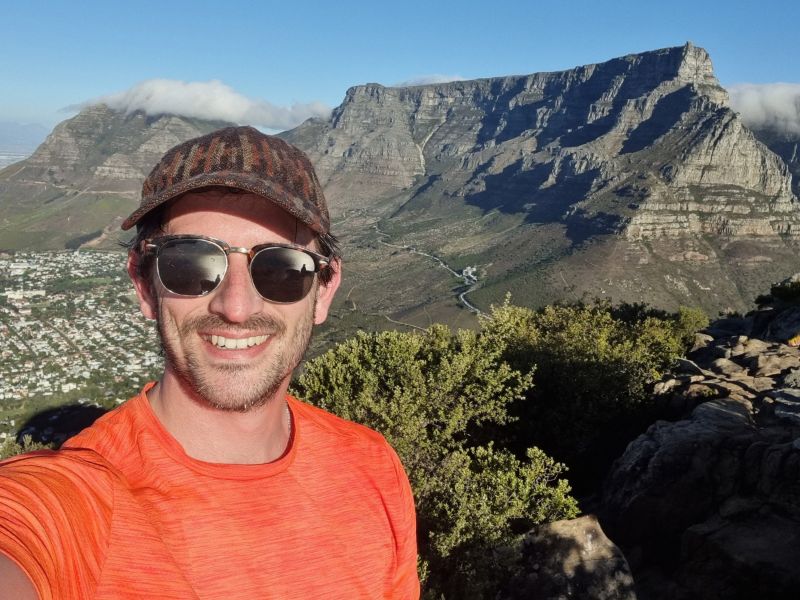 Dan smiling in an orange shirt and sunglasses, with Table Mountain behind him